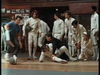 A group of people in fencing gear react as one fencer falls to the floor, while a person in casual clothes walks past holding a fencing helmet—capturing the kind of dramatic moment found in classic films like By The Sword DVD (1991). Others watch with concern or surprise.