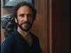 A man with curly hair and a beard stands indoors, wearing a dark shirt. Behind him, a dark sculpture or bust is set against a wooden wall, evoking the timeless ambiance of classic films like “By The Sword” (1991) on DVD.