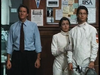 Three people stand indoors; one wears a shirt and tie, while two are dressed in white fencing uniforms—one holding a fencing mask and foil—resembling a scene straight from the “By The Sword DVD (1991).” Behind them, a bulletin board covered in papers adds to the authentic club atmosphere evocative of classic films like “By The Sword DVD (1991).”.