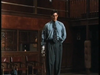 A man in a blue shirt and tie stands indoors holding a fencing sword, reminiscent of scenes from the “By The Sword DVD (1991).” His left arm appears to be a metallic prosthetic. The wooden walls, chairs, and balcony in the background evoke the unique atmosphere found in this rare film.