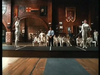 Two fencers in white uniforms face off on a raised platform in a wood-paneled room, while judges and spectators watch from the background—a striking scene reminiscent of By The Sword (1991) on DVD, capturing the elegance and intensity of traditional fencing duels.