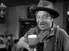 A man in a hat and jacket smiles while raising a glass of beer in a lively, old-fashioned bar reminiscent of a classic Western movie scene, as seen in the black-and-white film featured on the “20 Mule Team (1940) DVD.”.