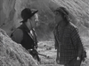 Two men stand in a rocky desert landscape straight out of a classic Western, captured in black and white on the “20 Mule Team (1940) DVD.” One wears a hat and vest while holding a rifle, as the other—dressed in a checkered shirt—faces him closely, appearing to argue.