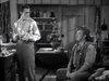 Two men in old-fashioned clothing are in a rustic kitchen; one stands by a table holding something, while the other sits on a chair, wearing a hat and smiling. Shelves and kitchen items appear in the background, capturing the charming Western atmosphere seen in classic films like “20 Mule Team (1940)”—now available on DVD.