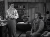Two men in vintage clothing converse inside a rustic kitchen; one stands by the counter holding an item, while the other, wearing a hat, sits and gazes up at him. With shelves and curtains in the background, the scene evokes the atmosphere of a classic Western—calling to mind moments from borax mining films such as 20 Mule Team (1940) DVD.