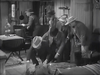 Six people in cowboy hats gather around another person lying on the floor in a rustic room, while one person sits at a table in the background. The black and white scene exudes an old Western atmosphere, reminiscent of classic films like “20 Mule Team (1940)”, available on DVD.