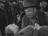 A man in a hat urgently speaks to a woman lying down with her eyes closed, surrounded by concerned onlookers in a crowded, black-and-white scene from the classic Western film “20 Mule Team (1940),” as featured on DVD.