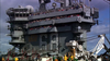 A large aircraft carrier, bustling with crowds across multiple decks, showcases crew members in colored uniforms working on the deck below amid visible equipment and machinery. This striking scene recalls memorable moments from a military family film such as the Tiger Cruise (2004) DVD, capturing the excitement and camaraderie found aboard a naval ship.