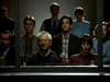 A group of serious-looking adults sit closely together in a dimly lit room, facing forward, as seen through a window. Some wear glasses; their expressions are attentive and somber, as if gathered to unravel an enigma worthy of “Evidence In Blood DVD (1998),” a rare movie collectible.