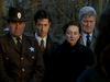 Four somber adults, including a uniformed officer and three others in dark formal clothing, stand close together outdoors, appearing solemn as they attend a ceremony or gathering. Leafless trees fill the background, creating an eerie atmosphere reminiscent of scenes from the Evidence In Blood DVD (1998).