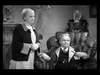 An elderly woman stands beside an older man seated in a large armchair. Both appear serious in a dimly lit, vintage room with ornate furnishings—like characters captured from Make Way For Tomorrow DVD (1937).