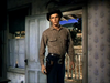 A young man wearing a cowboy hat and checkered shirt stands in a doorway, gun holster at his side, peering into a rustic house with a porch and rural backdrop—evoking the timeless spirit of classic westerns like Gun Belt (1953) DVD.