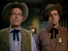 Two men dressed in cowboy hats, sheriff badges, and bow ties stand indoors, channeling the spirit of classic westerns—much like the cover of the Gun Belt (1953) DVD. One sports a tan vest over a white shirt, while the other wears a brown shirt. The dimly lit background with a lantern on the wall further evokes the timeless atmosphere found in cowboy cinema.