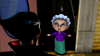 An animated elderly woman with glasses, gray hair, a purple sweater, and a green skirt stands in a doorway, smiling with arms open—as if welcoming someone to enjoy a Halloween movie night or discover rare movies on DVD such as Dear Dracula (2012) DVD—facing a person in the foreground.