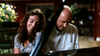 A smiling woman and a man sit closely together at a piano, both looking down. The softly lit background with a flower arrangement on the left evokes the timeless charm of classic films like “Love, Murder and Deceit (1997)” on DVD.