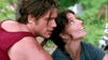 A man in a red tank top leans close to a smiling woman with curly hair, who is looking upward. They appear to be outdoors with greenery in the background, sharing a joyful moment reminiscent of scenes from “Love, Murder and Deceit (1997)” on DVD.