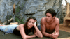 A woman in patterned leggings and a man, both lying on their stomachs on a mat outdoors near a rocky wall, relax and smile quietly together, perhaps discussing their favorite rare DVDs like “Love, Murder and Deceit (1997).”.