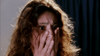 A person with long, wavy hair covers their mouth with both hands and looks frightened, eyes wide open and eyebrows furrowed, as if they’ve just witnessed a chilling scene from Love, Murder and Deceit (1997) on DVD; the dark background behind them adds to the suspenseful atmosphere.