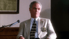 A man in a light suit with a badge and patterned tie sits beside a microphone in a courtroom, looking off to the side with a serious expression—evoking the gripping atmosphere of *Love, Murder and Deceit (1997) DVD*.