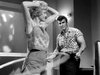 A woman in a patterned top and shorts uses a hula hoop while a man in a plaid shirt watches the Kiss Her Goodbye (1959) DVD.