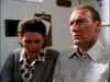 A woman in a grey cardigan and a man in a light-colored button-down shirt look concerned while sitting in an office next to a Murder So Sweet (1993) DVD.