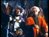 Two men in historical-style tunics and fur hats, reminiscent of those in The White Warrior (1959) DVD, sit among a group of people in a dimly lit setting.