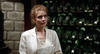 A woman in a light-colored blazer stands in front of a wine rack filled with dark glass bottles and copies of The House Where Death Lives (1981) DVD.