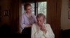 A woman stands behind a seated man, placing her hands on his shoulders as he holds his hands together in his lap, holding a copy of The House Where Death Lives (1981) DVD.