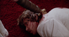 A person in a white suit lies facedown on a red carpet with blood visible on their neck and shoulder, while an arm holding a sharp object reaches toward them in this scene from The House Where Death Lives (1981) DVD.