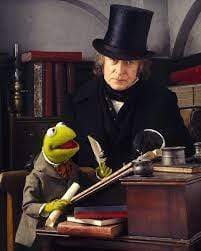 Kermit the Frog in Victorian attire sits with Michael Caine amid books and writing tools—a scene from The Muppet Christmas Carol, featured in The Muppet Christmas Carol DVD (1992).