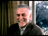 An older man with short gray hair and a beard smiles by a window framed by trees, wearing a brown jacket over a collared shirt, perhaps reminiscing about classic films like the "Amos (1985) DVD.