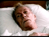 An elderly man with gray hair lies in a hospital bed, looking weak and tired—his hand on his chest—as the senior care drama "Amos (1985) DVD" quietly unfolds.