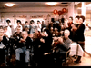 A group of elderly people and staff celebrate in a decorated room under a "Merry Christmas" banner, smiling and clapping at a holiday event in a care facility featuring the Amos (1985) DVD.