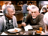 Two older men sit at a cafeteria table eating breakfast, with plates of food and orange juice—reminiscent of a scene from a senior care drama like the one in "Amos (1985)" DVD. Other people appear in the bustling background.